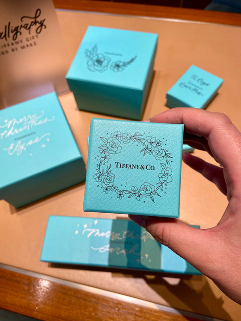 A hand holds a Tiffany & Co. box which has been customized with hand drawn florals. Other customized blue boxes sit further on a desk. 