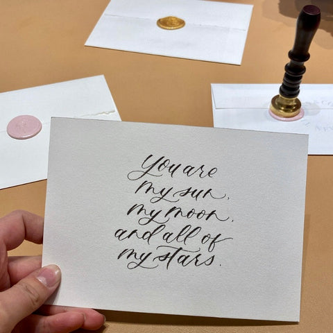 You are my sun, my moon and all of my stars written in modern calligraphy on a note card for Tiffany & Co. Behind the note there are envelopes with gold and pink wax seals are scattered on a desk in the background.