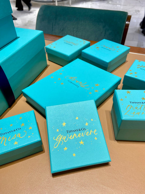 Multiple blue Tiffany & Co. boxes lay on a desk. Each box is customized by Chalked by Mabz. The boxes are decorated with gold calligraphy and stars