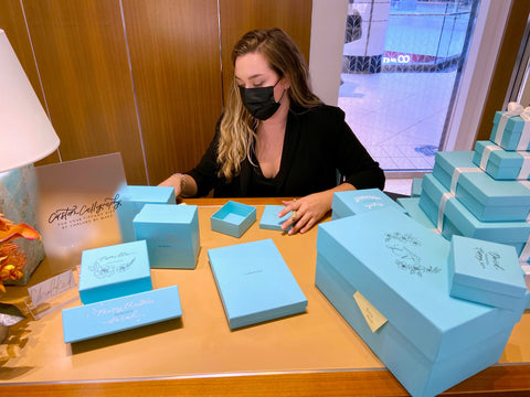 Modern calligrapher Chalked by Mabz sits behind a desk. The desk is covered in Tiffany blue boxes. Each box is uniquely decorated with floral illustrations and calligraphy. 
